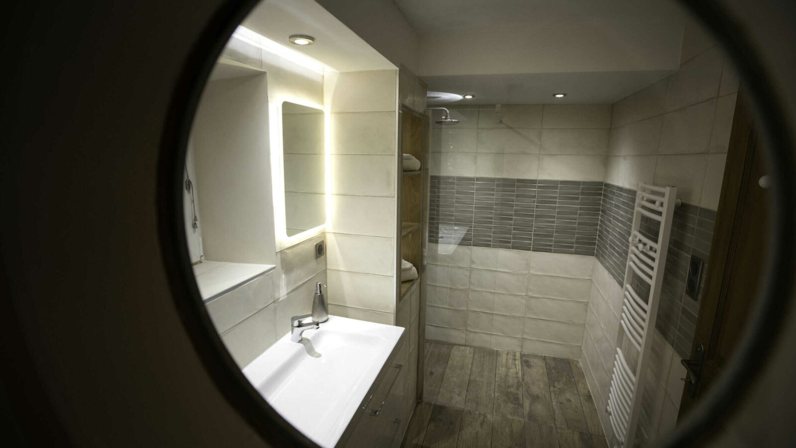 View through a round window showing a modern bathroom with sink, mirror, towel radiator, tiled walls, wooden floor, and walk-in shower with glass partition at Mas Saint Antoine gîte Ardèche. Enjoy comfort just steps from the piscine. View through a round window showing a modern bathroom with sink, mirror, towel radiator, tiled walls, wooden floor, and walk-in shower with glass partition at Mas Saint Antoine gîte Ardèche. Enjoy comfort just steps from the piscine.