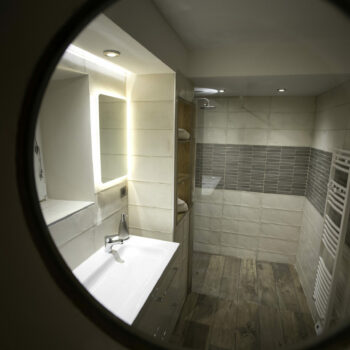 View through a round window showing a modern bathroom with sink, mirror, towel radiator, tiled walls, wooden floor, and walk-in shower with glass partition at Mas Saint Antoine gîte Ardèche. Enjoy comfort just steps from the piscine.