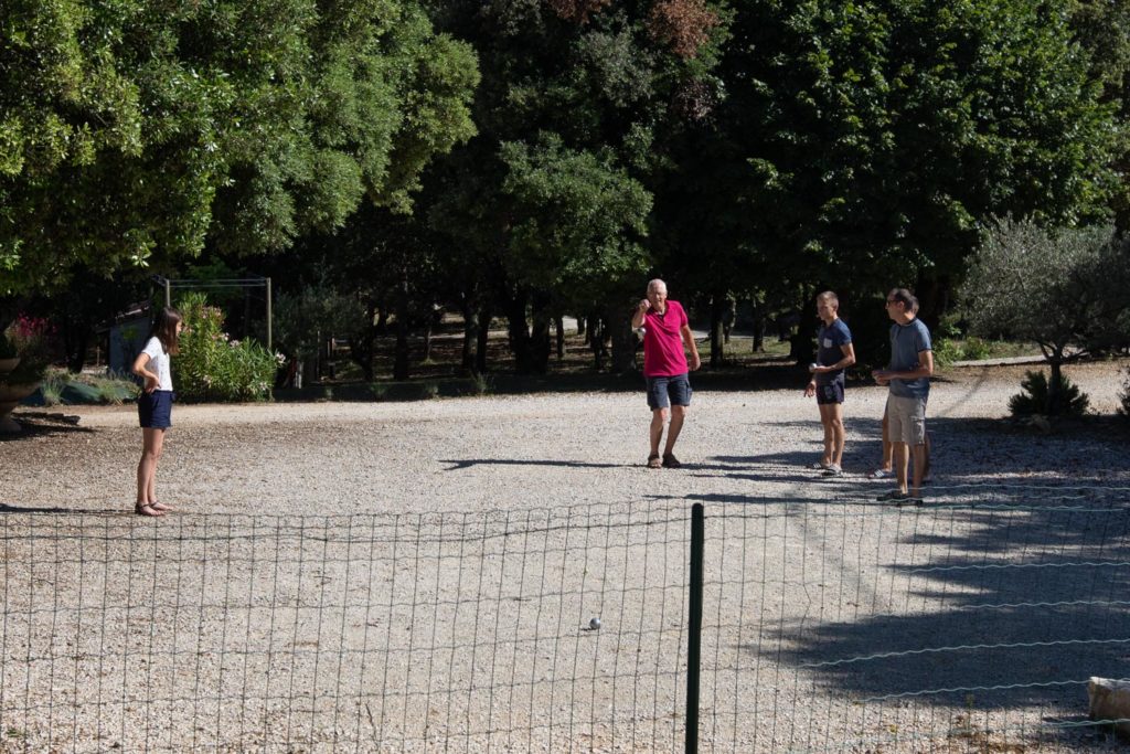 Quatre personnes se tiennent en plein air sur un terrain en gravier entour&eacute; d'arbres au Mas Saint Antoine, avec un grillage au premier plan. Trois personnes sont ensemble &agrave; droite, une personne est seule &agrave; gauche et la lumi&egrave;re du soleil passe &agrave; travers. C'est l'endroit id&eacute;al pour votre h&eacute;bergement Ard&egrave;che m&eacute;ridionale.