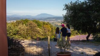 Trois personnes, deux enfants et un adulte au chapeau rose, se tiennent sur un chemin caillouteux surplombant des montagnes, de la verdure, un champ de lavande et un gîte ardéchois sous un ciel bleu limpide.