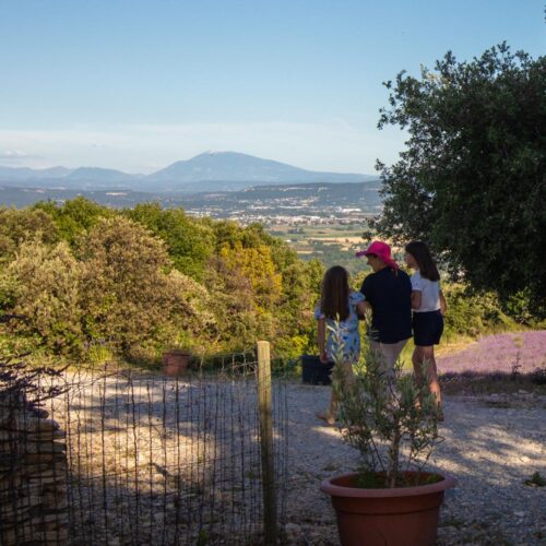 Trois personnes, deux enfants et un adulte au chapeau rose, se tiennent sur un chemin caillouteux surplombant des montagnes, de la verdure, un champ de lavande et un gîte ardéchois sous un ciel bleu limpide.