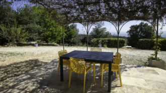 Un patio avec une table noire et quatre chaises jaunes se trouve sous une pergola couverte de vignes dans ce gîte de l'Ardèche, avec vue sur une cour en gravier et des arbres verts en arrière-plan lors d'une journée ensoleillée.