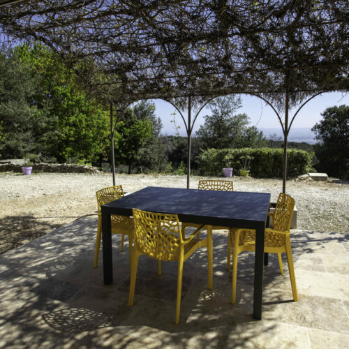 Un patio avec une table noire et quatre chaises jaunes se trouve sous une pergola couverte de vignes dans ce gîte de l'Ardèche, avec vue sur une cour en gravier et des arbres verts en arrière-plan lors d'une journée ensoleillée.