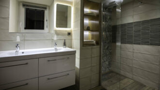 Salle de bain moderne au Mas Saint Antoine avec un double lavabo blanc, un miroir éclairé, une petite fenêtre, des étagères en bois ouvertes avec des serviettes, et une douche vitrée avec des carreaux à motifs et gris - parfait pour votre séjour dans un gîte en Ardèche.
