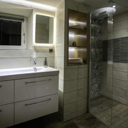 Salle de bain moderne au Mas Saint Antoine avec un double lavabo blanc, un miroir éclairé, une petite fenêtre, des étagères en bois ouvertes avec des serviettes, et une douche vitrée avec des carreaux à motifs et gris - parfait pour votre séjour dans un gîte en Ardèche.