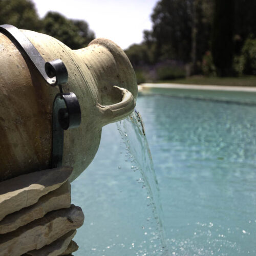 Une cruche rustique en céramique verse de l'eau dans une piscine turquoise au Mas Saint Antoine, un hébergement de charme en Ardèche méridionale pour les séjours en groupe, entouré de bordures en pierre et d'une verdure luxuriante sous un soleil éclatant.