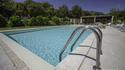 Une piscine claire avec des rampes en métal, des dalles en pierre, des chaises longues et des arbres sous un ciel bleu vif dans un gîte de l'Ardèche ; un bâtiment au bord de la piscine est visible à l'arrière-plan.