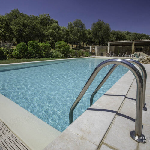 Une piscine claire avec des rampes en métal, des dalles en pierre, des chaises longues et des arbres sous un ciel bleu vif dans un gîte de l'Ardèche ; un bâtiment au bord de la piscine est visible à l'arrière-plan.