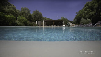 Une piscine claire entourée de chaises longues et de verdure, avec un patio ombragé en arrière-plan sous un ciel bleu, invite à la détente au Mas Saint Antoine, votre hébergement idéal Ardèche méridionale.