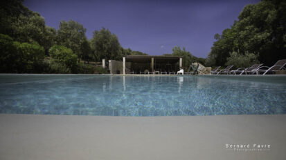 Une piscine claire entourée de chaises longues et de verdure, avec un patio ombragé en arrière-plan sous un ciel bleu, invite à la détente au Mas Saint Antoine, votre hébergement idéal Ardèche méridionale.