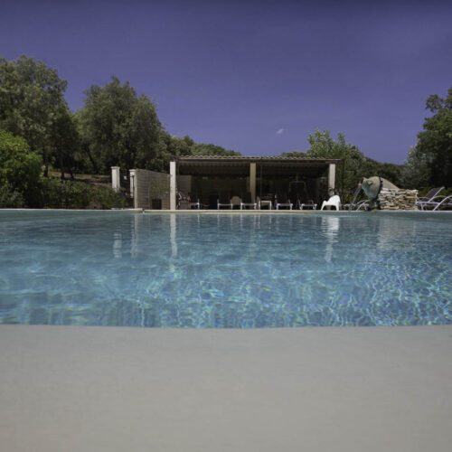 Une piscine claire entourée de chaises longues et de verdure, avec un patio ombragé en arrière-plan sous un ciel bleu, invite à la détente au Mas Saint Antoine, votre hébergement idéal Ardèche méridionale.