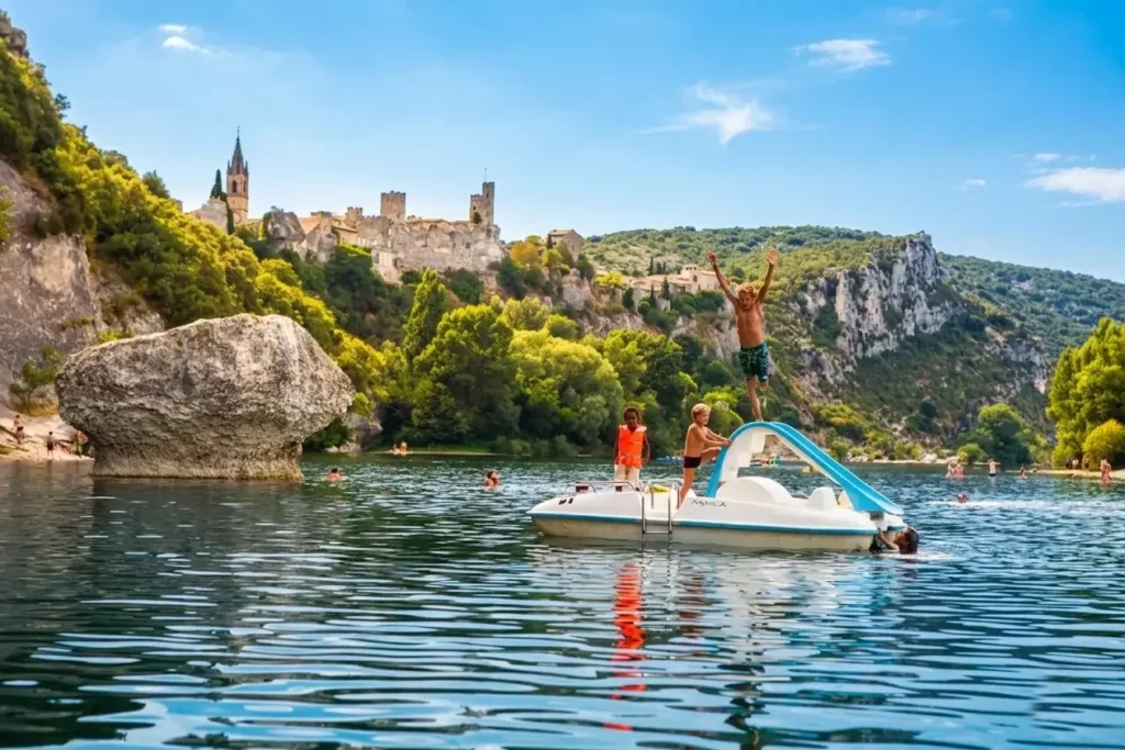People are enjoying a paddleboat with a slide on a calm river near rocky cliffs and lush vegetation; one person is jumping off the slide while others watch. A historic hilltop village and the Mas Saint Antoine, a charming Ard&egrave;che guesthouse, are visible in the background.